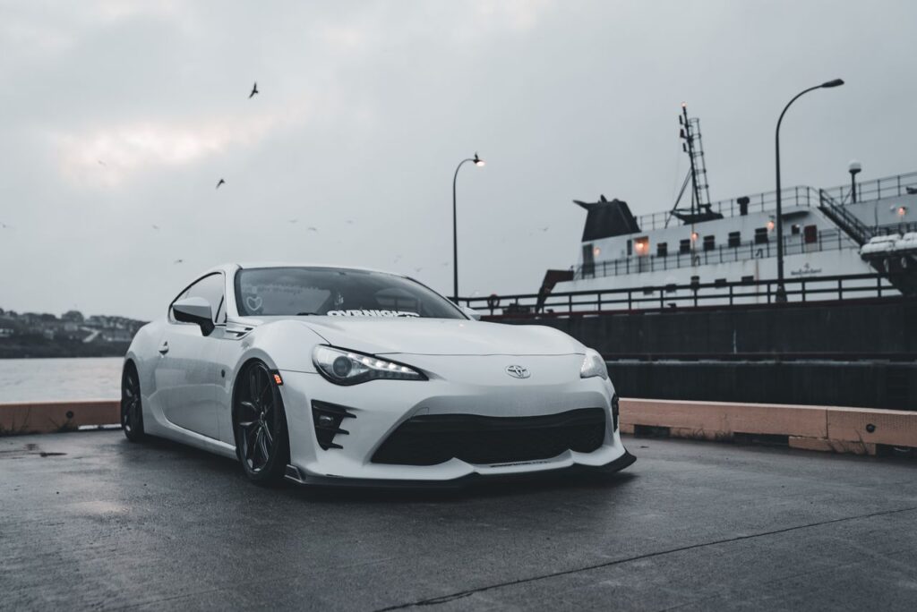 White sports car parked near a harbor with a ferry in the background at dusk, creating a moody scene.