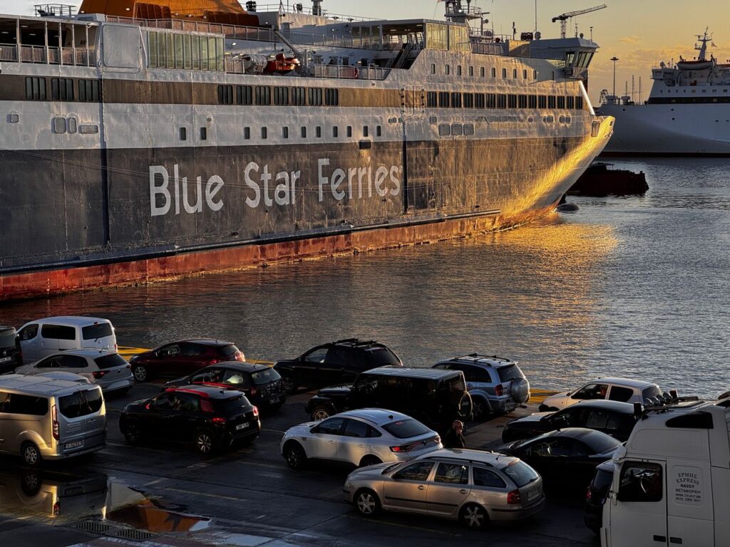 A Blue Star Ferry docked at Athens port during sunset with cars parked nearby.
