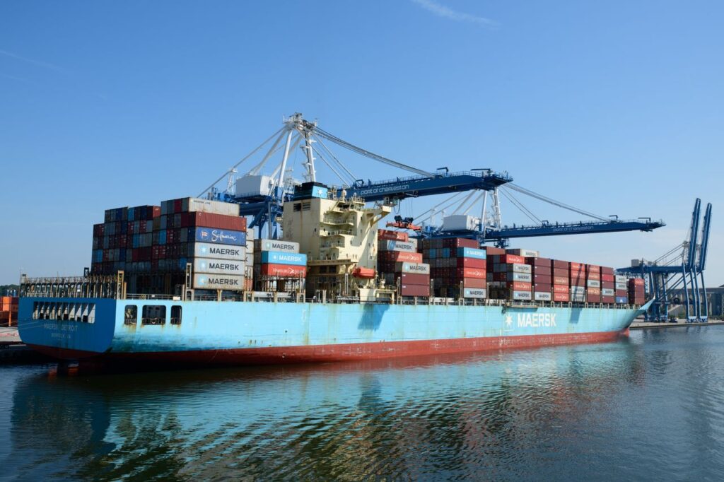 A large cargo ship loaded with containers at a busy port under a clear blue sky.